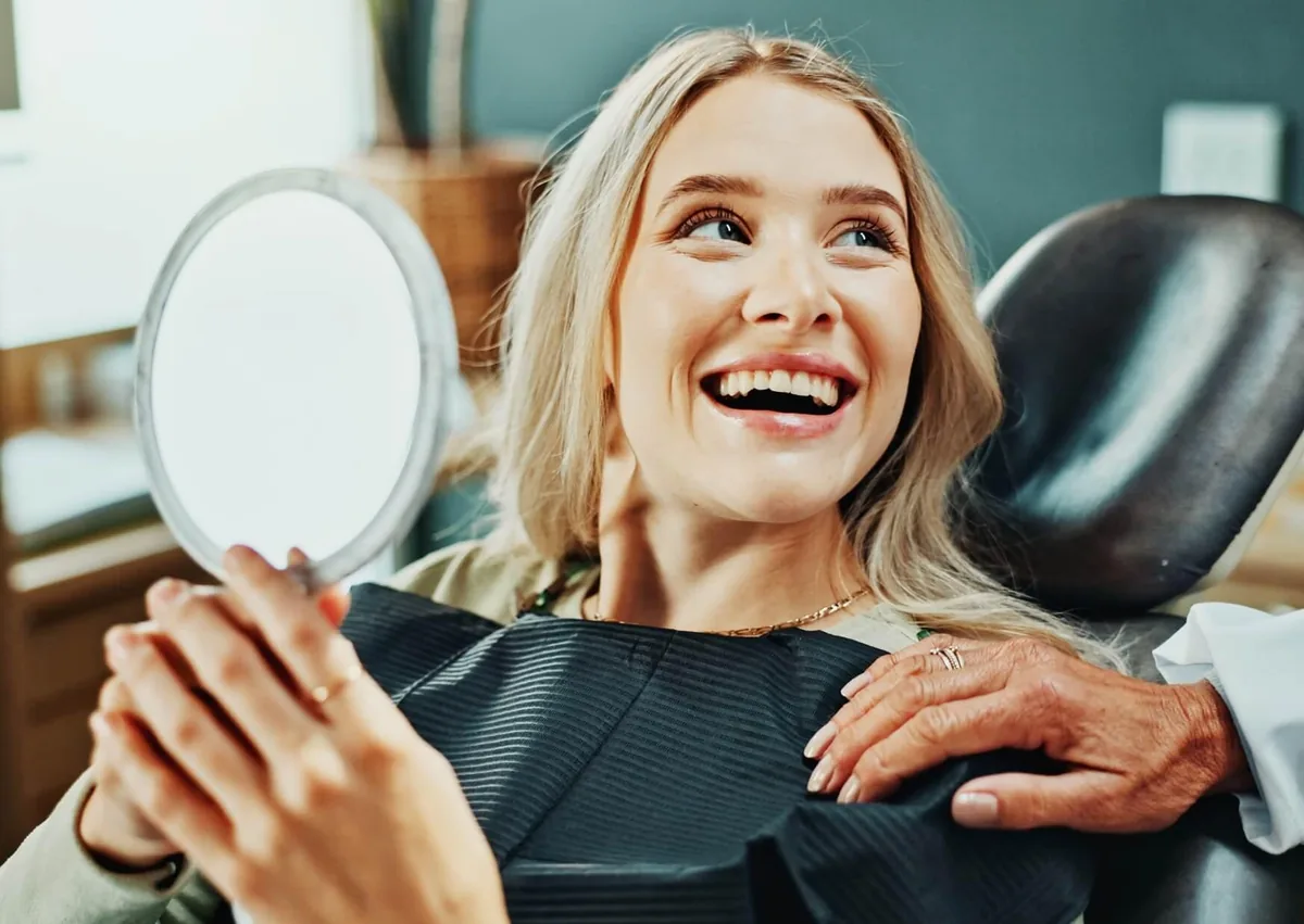 Smiling patient holding a mirror to view her teeth during a digital smile design consultation at a dental clinic.